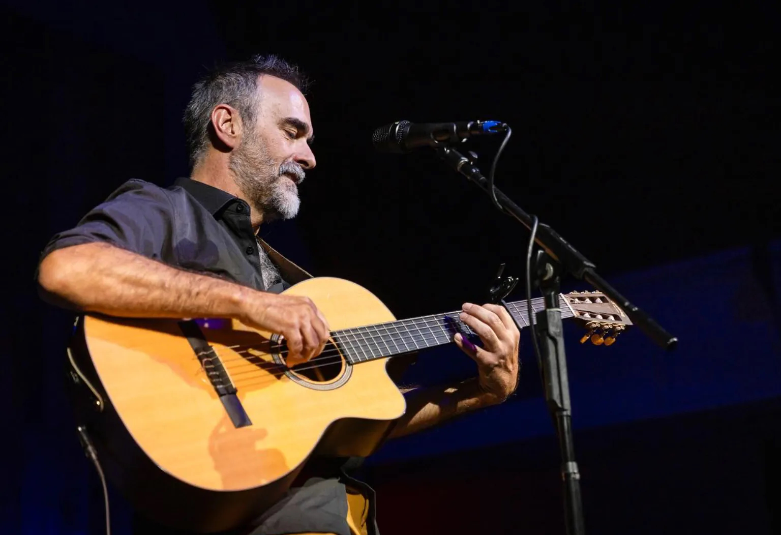 Man playing acoustic guitar on stage with microphone, dark background, focused expression.