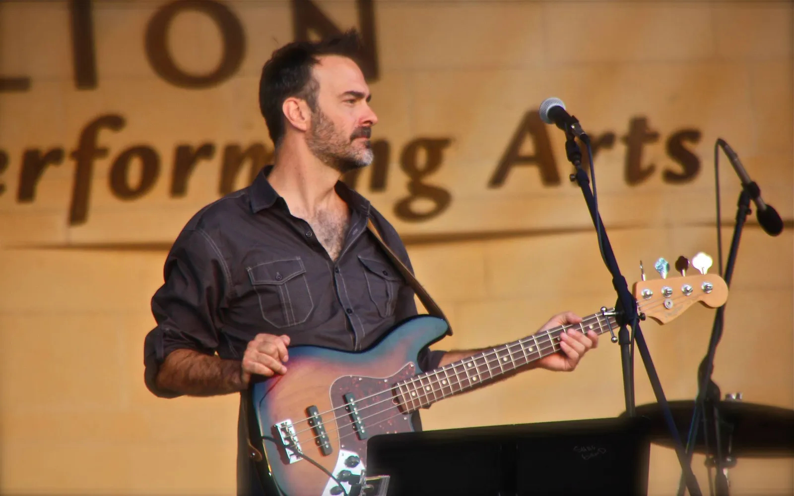 Man playing bass guitar on stage with microphones and a banner in the background.