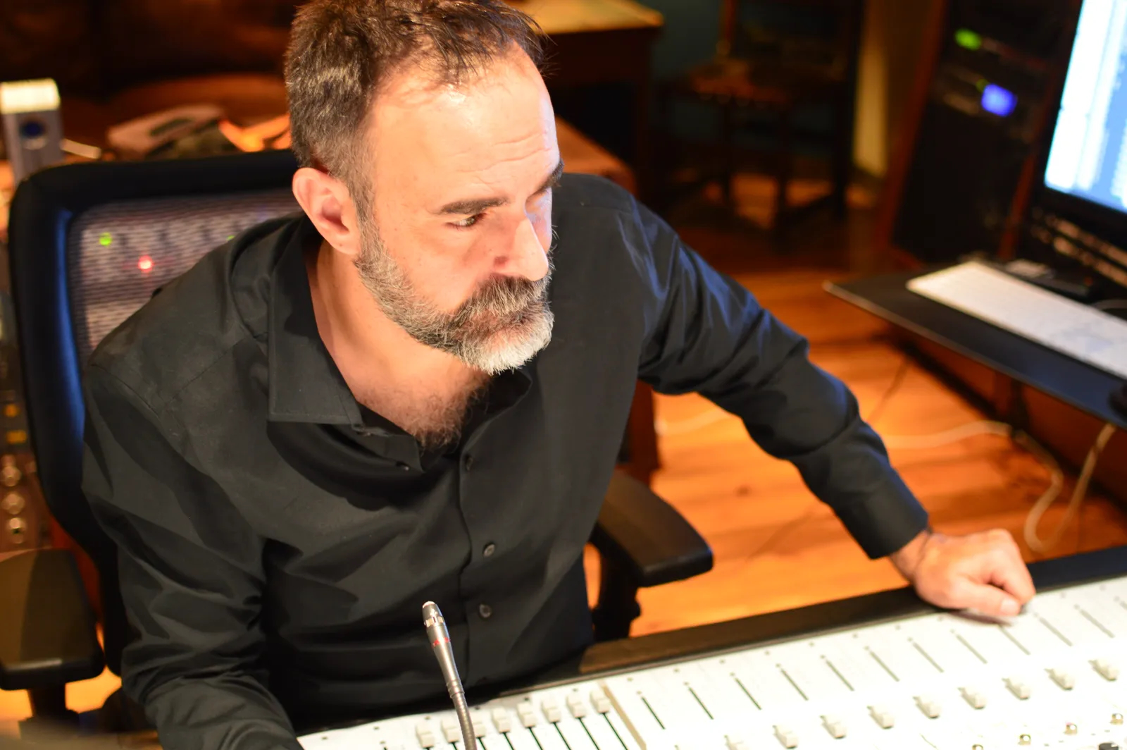 Man with beard and dark shirt sitting at a desk with audio equipment, looking to the side.