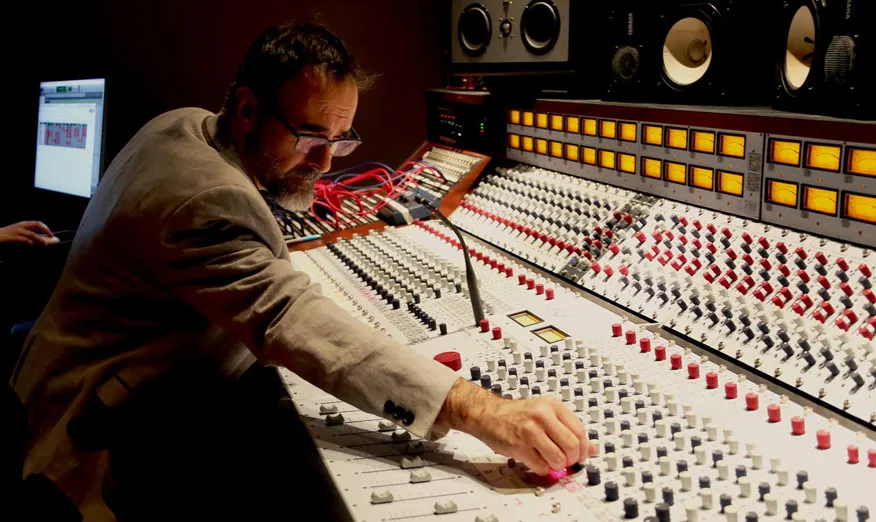 Person adjusting audio mixing console in a recording studio with equipment and speakers.