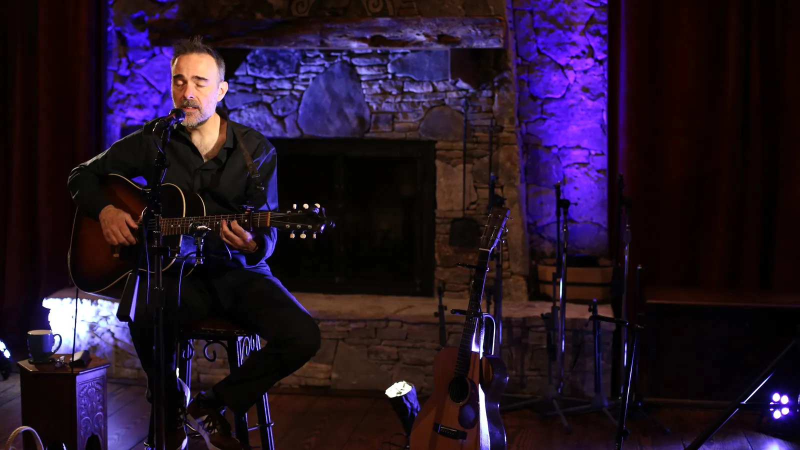 Man playing guitar on stage with purple lighting and a brick fireplace in the background.