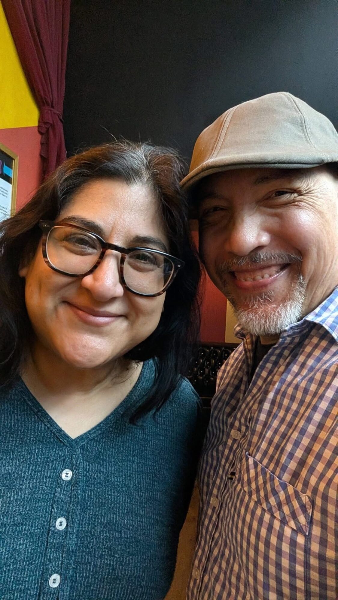 Two people smiling, woman with glasses and dark hair, man with a hat and beard, in an indoor setting.