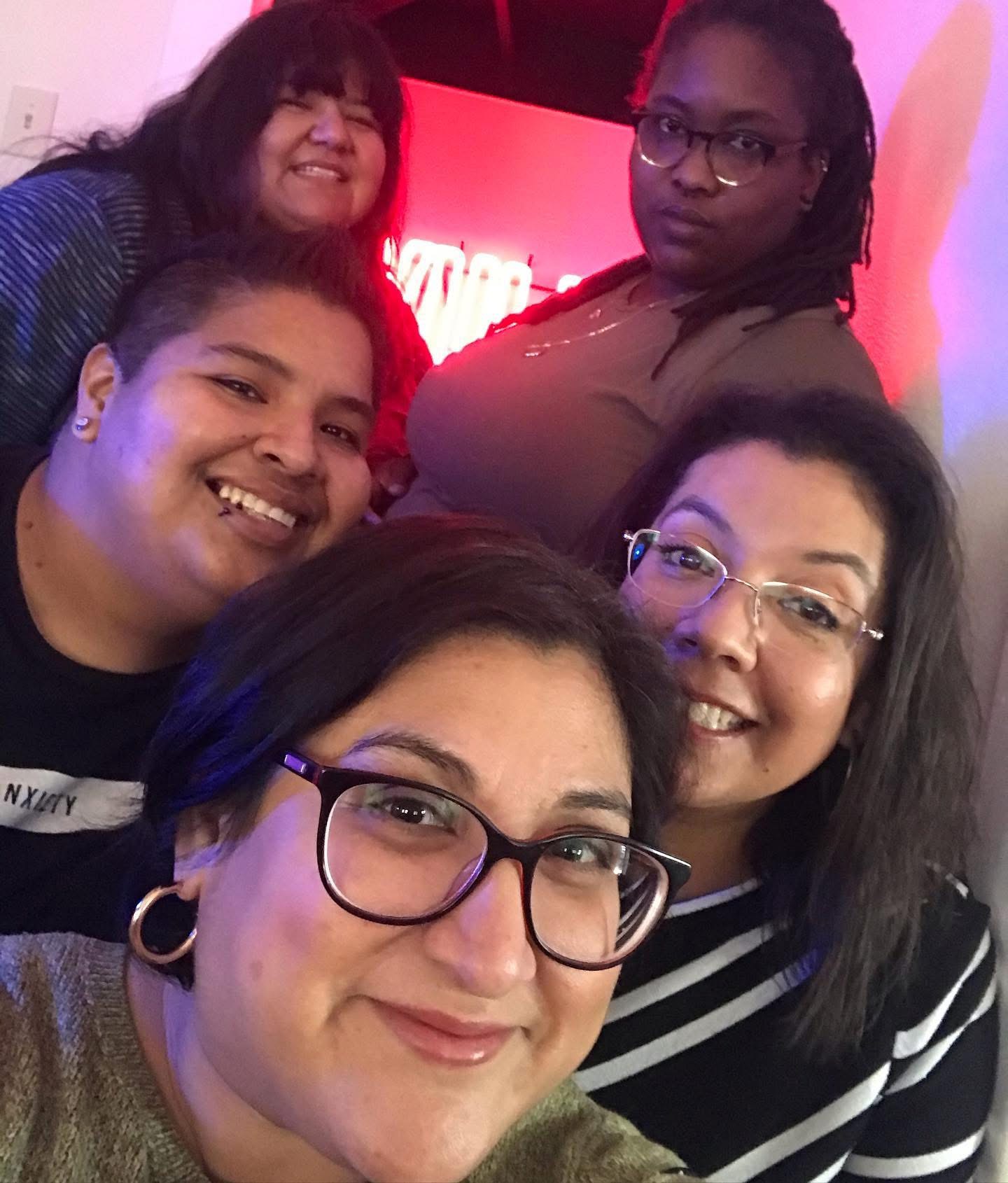 Five women smiling and posing for a group selfie indoors, with a red background and a screen behind them.