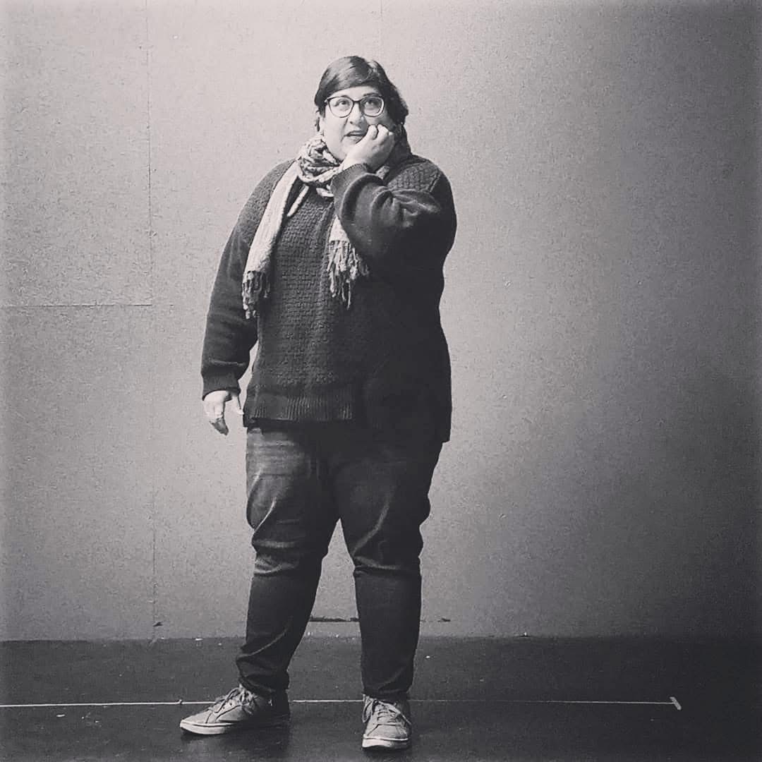 Person standing against a plain wall, wearing glasses, a scarf, and casual clothing, with one hand near their face.