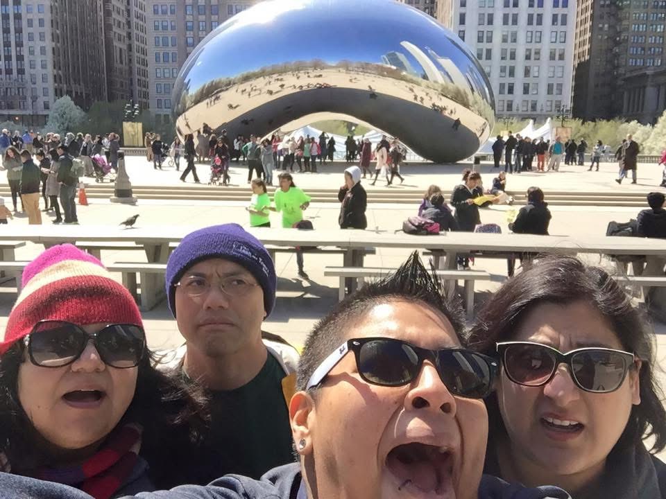 Four people taking a selfie in front of Cloud Gate sculpture in Millennium Park, Chicago, with a cityscape background.