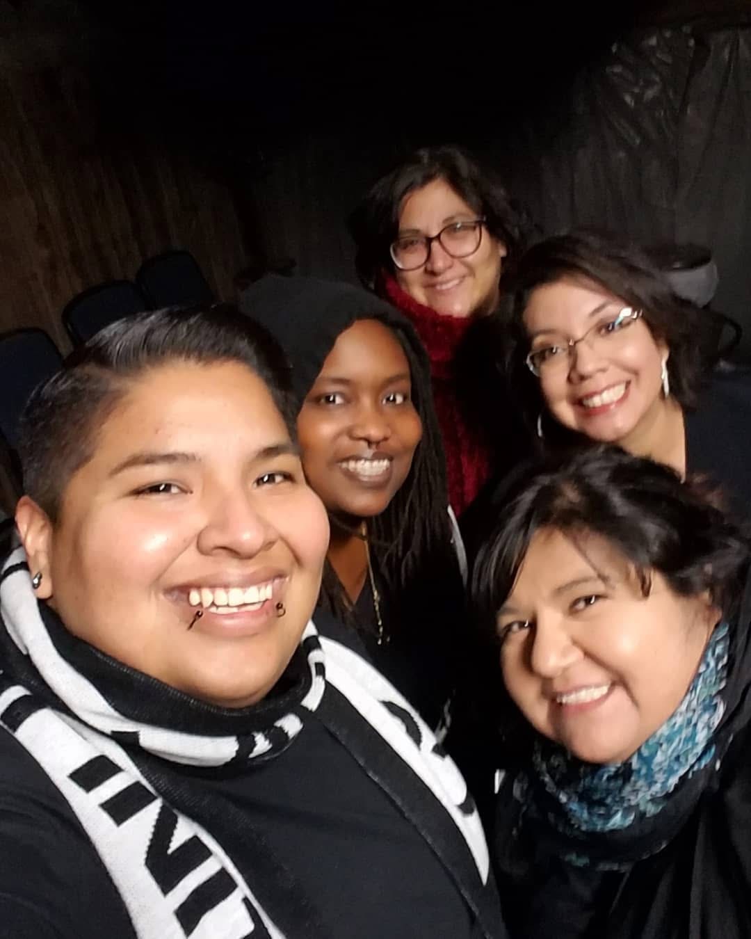 Group of five women smiling together indoors, some wearing glasses, with dark background.