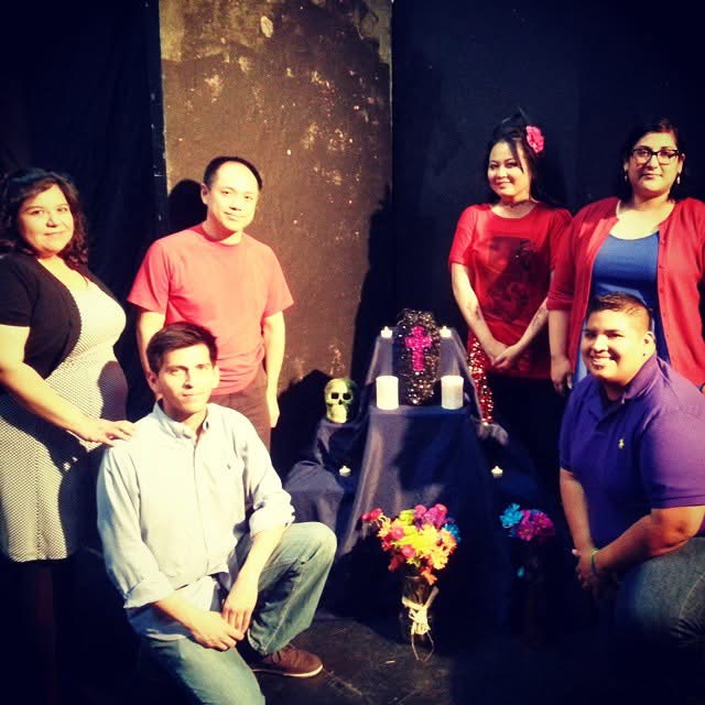 Group of six people standing around a table with a skull, candles, and flowers, in a dimly lit setting.