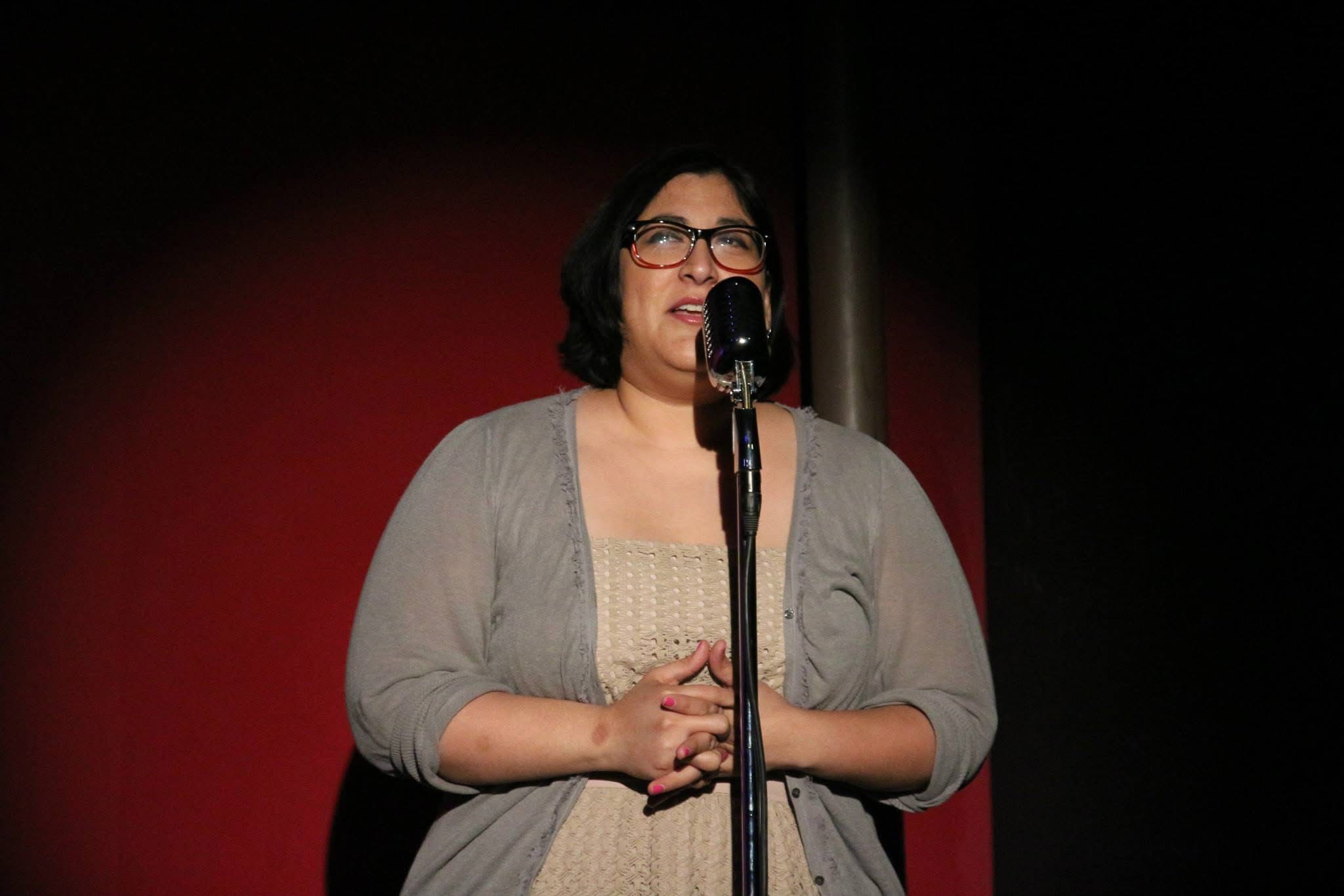 Woman with glasses speaking into a microphone on stage, hands clasped, background is red and black, indoor setting.