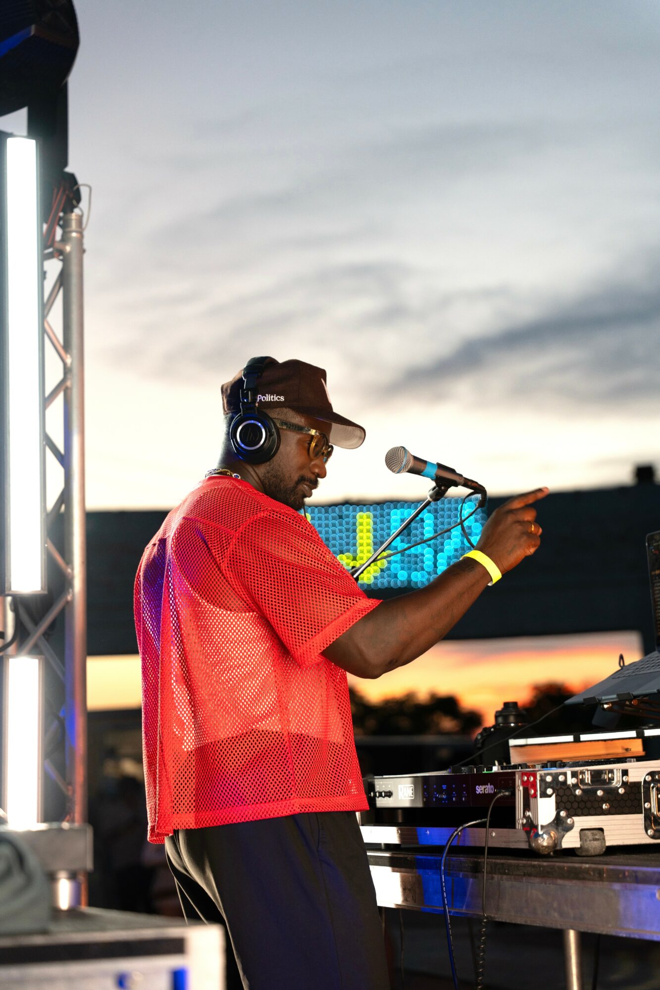 DJ wearing headphones and a red shirt, pointing at equipment during outdoor event at sunset.