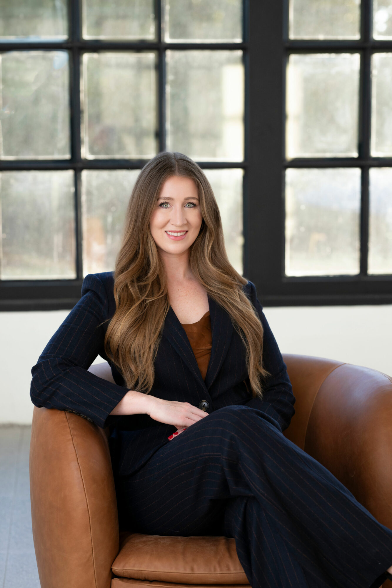 Woman with long hair sitting on a brown leather chair in front of large window, smiling, wearing a dark blazer and pants.