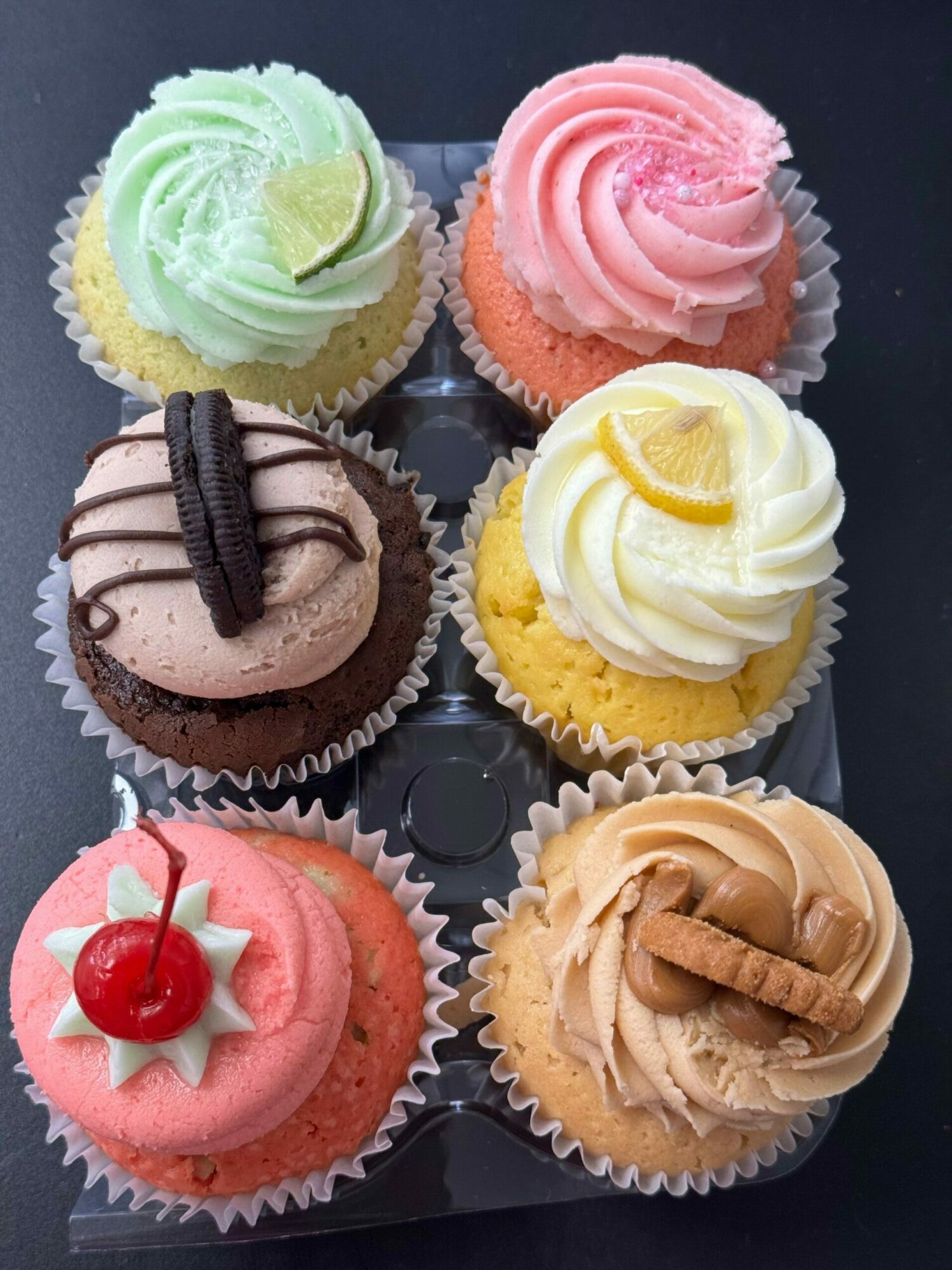 Six cupcakes with colorful frosting and toppings arranged in two rows, on a black surface.