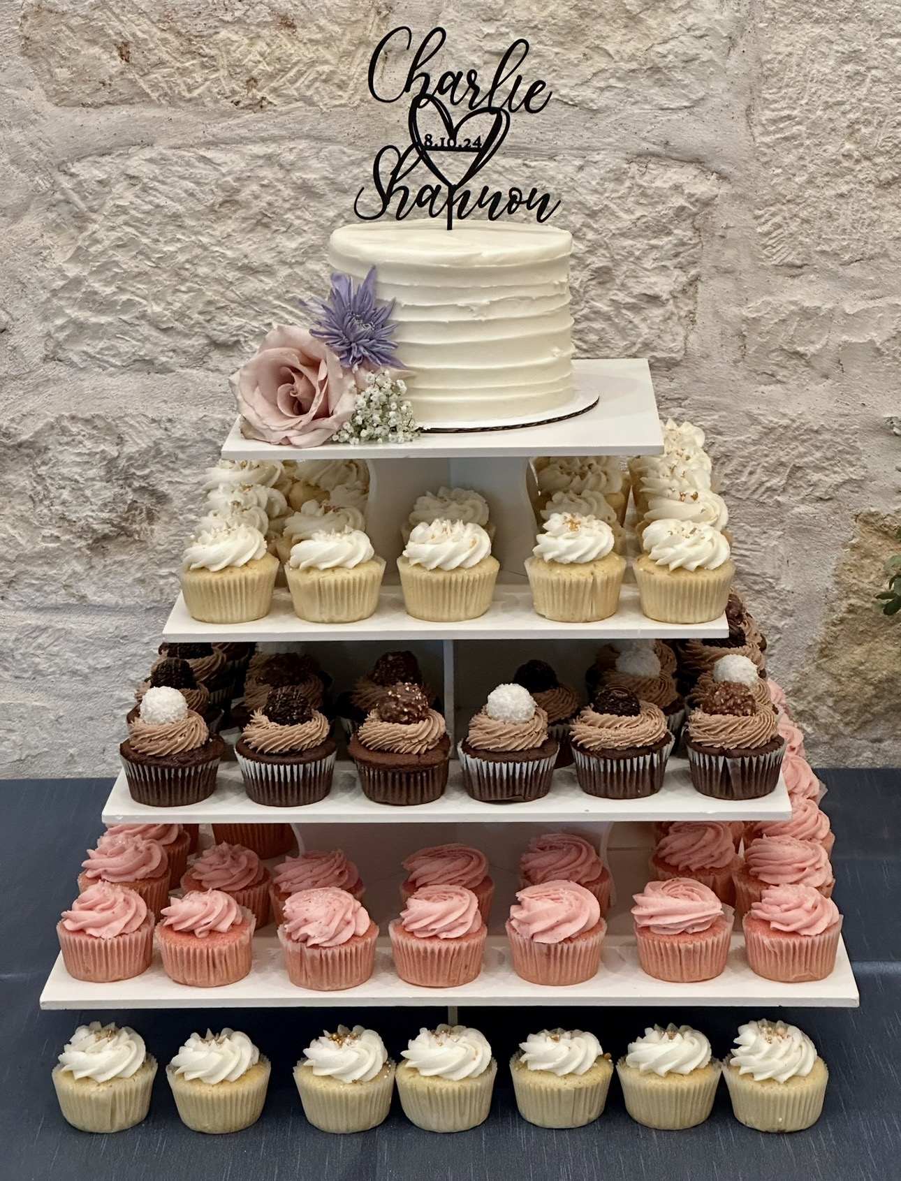 Tiered cake with cupcakes, decorated with flowers and a cake topper, set against a stone wall background.
