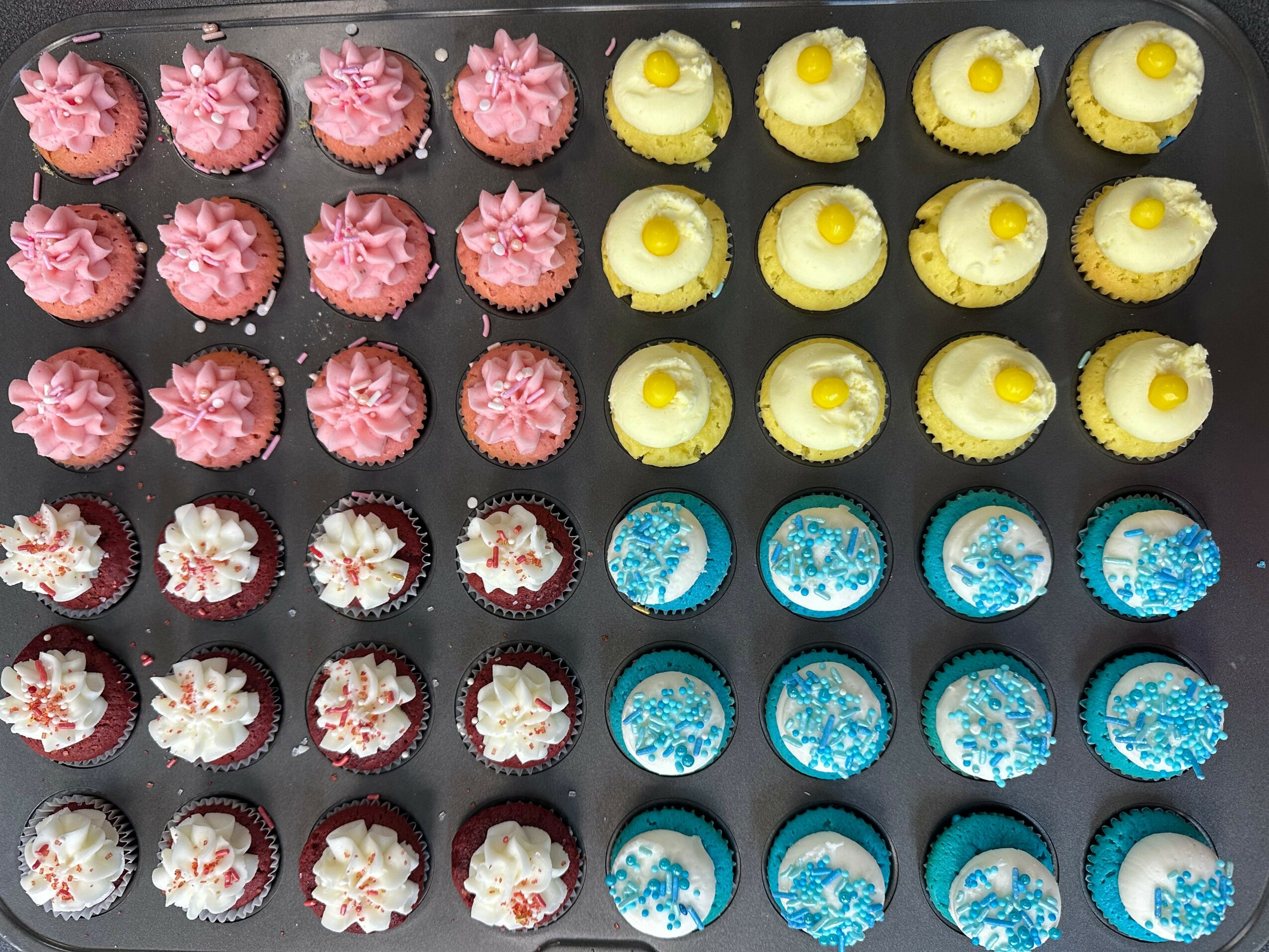 Assorted cupcakes with pink, yellow, white, and blue frosting, arranged in rows on a black tray.