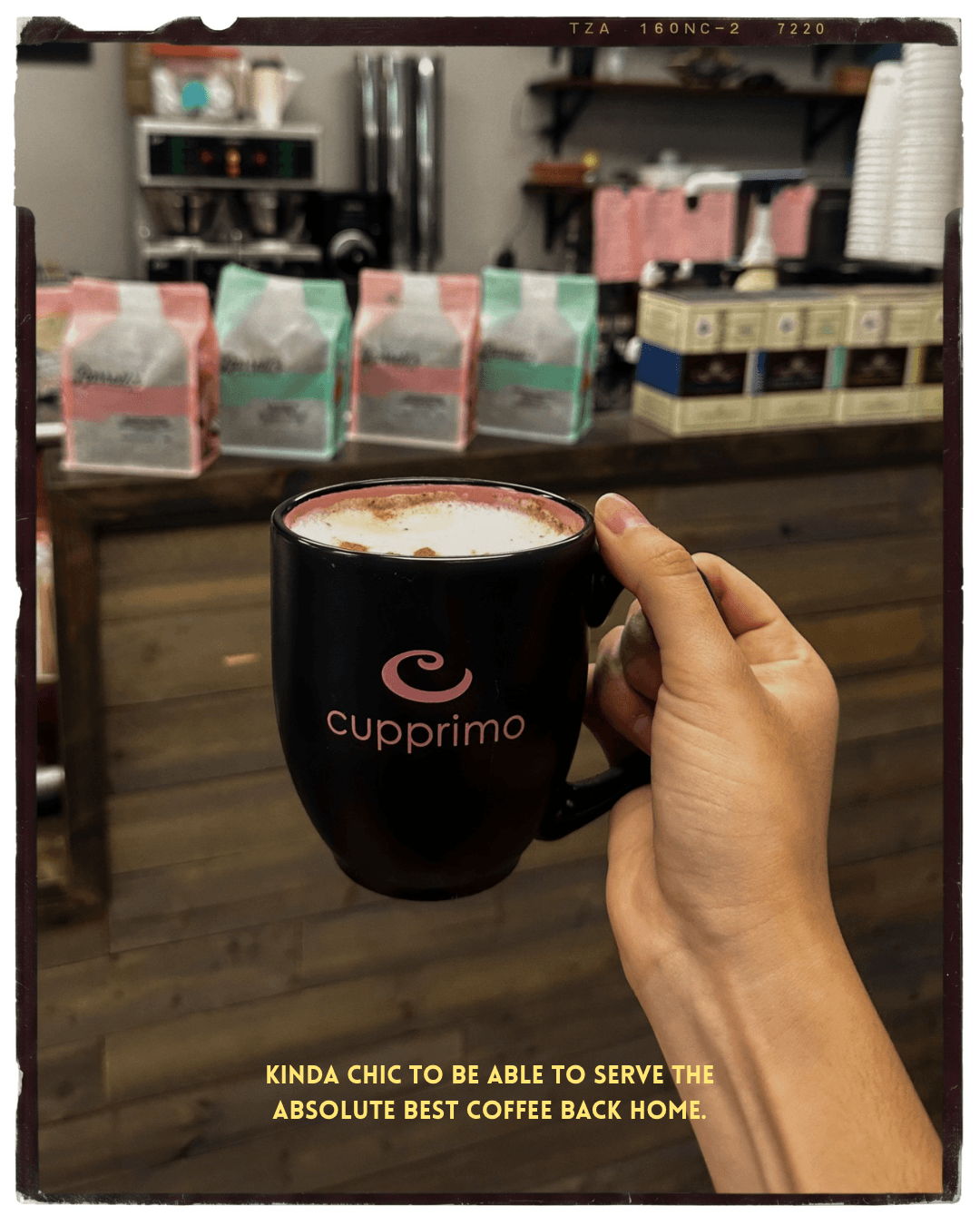 Hand holding a black coffee mug with pink logo, coffee inside, in a cafe setting with boxes and shelves in background.