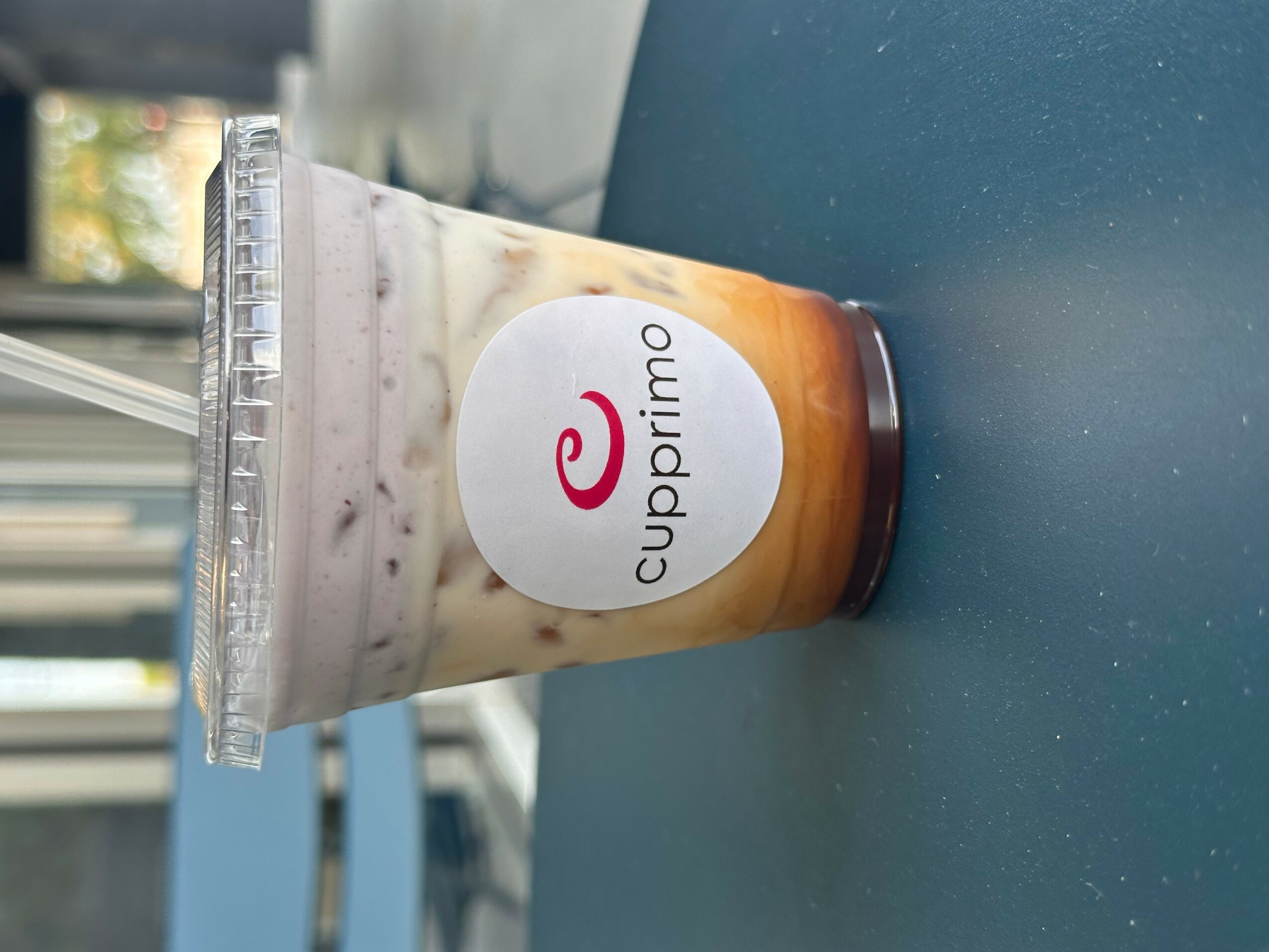 Cupprimo iced coffee in a clear plastic cup with a white label and a red logo, on a blue surface.