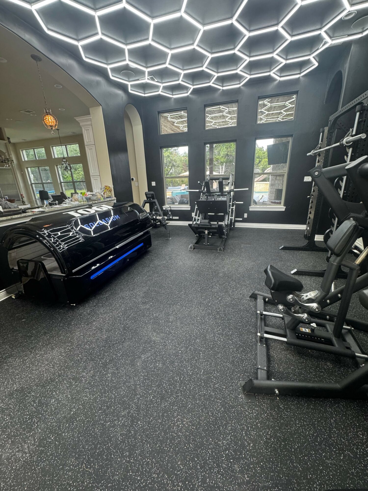 Gym with hexagonal ceiling lights, exercise equipment, and large windows, black rubber flooring, and a black car in the corner.