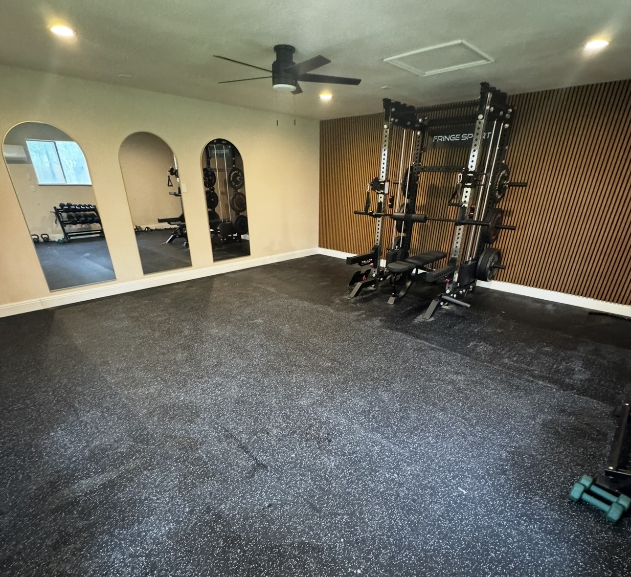 Empty gym room with black rubber flooring, a power rack, and mirrors on the wall, ceiling fan, and wooden wall paneling.