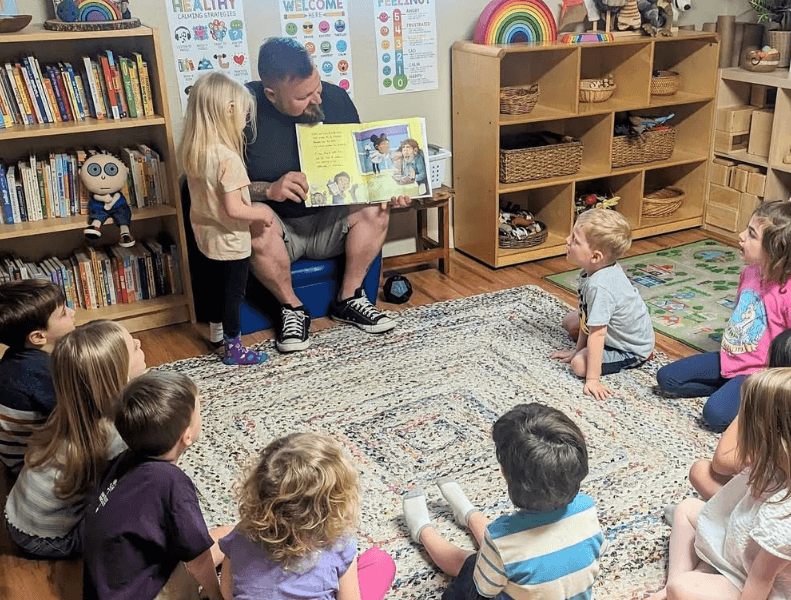 Adult reading a children's book to a group of children sitting on a colorful rug in a classroom.