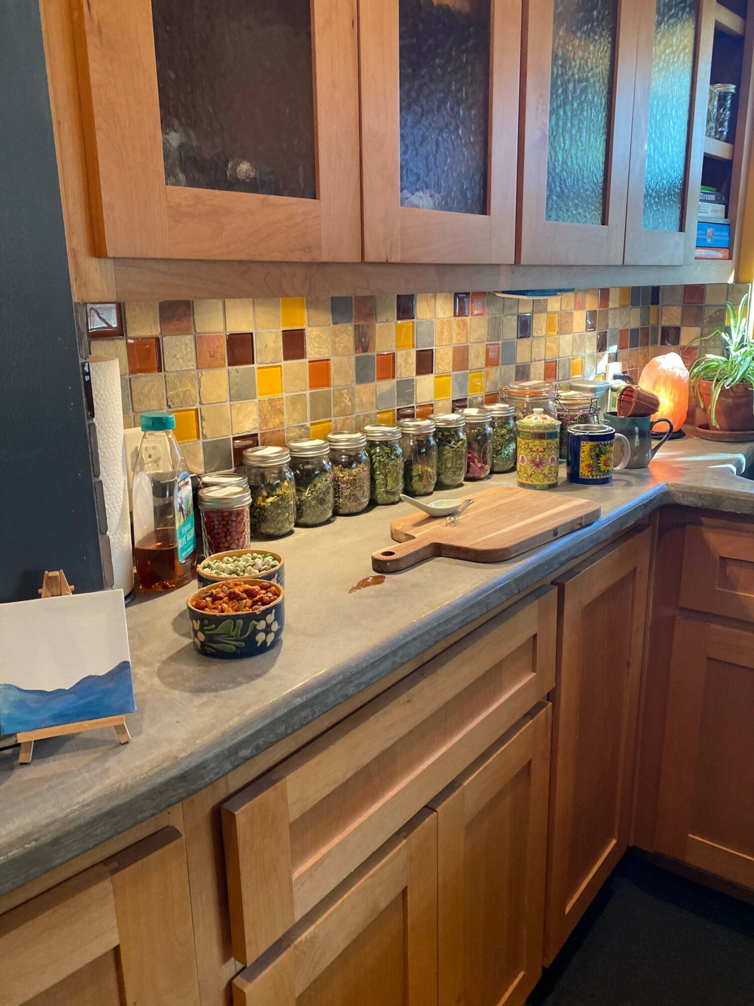 Kitchen counter with spice jars, a small container, and a cutting board, with wooden cabinets and a tiled backsplash.
