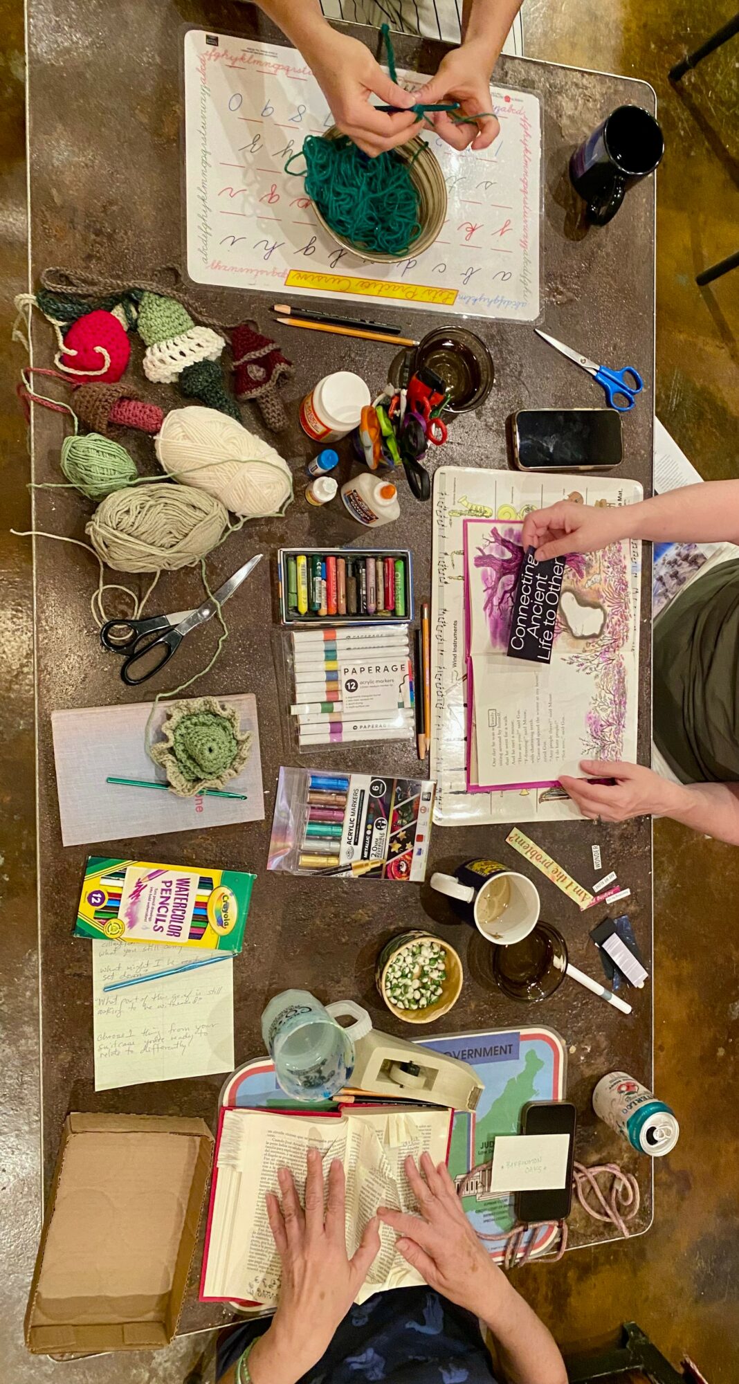 People working on knitting and crochet projects with yarn, needles, and tools on a table, viewed from above.