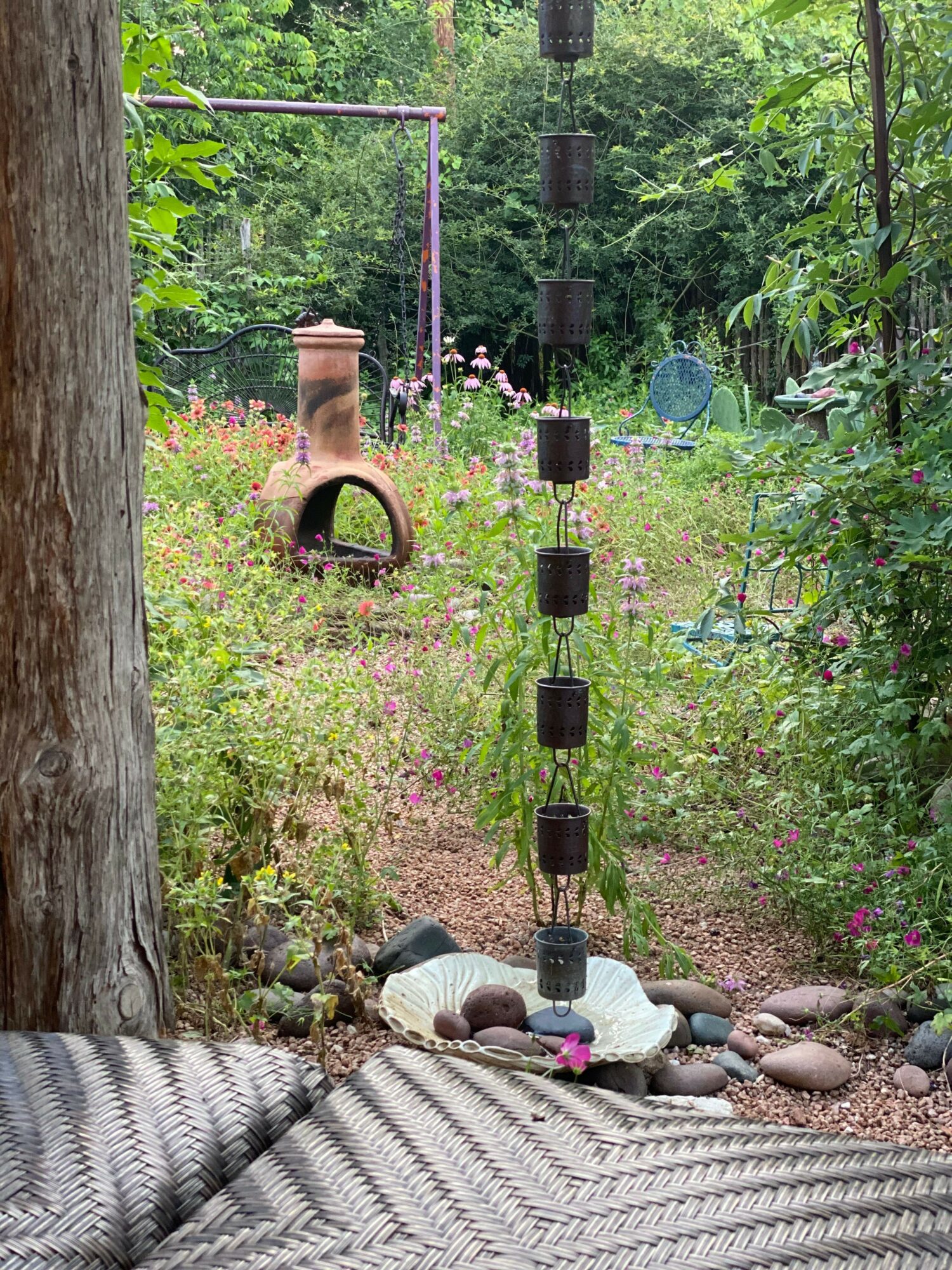 Garden scene with a watering can, plants, and a tree trunk, viewed from a side angle with outdoor furniture.