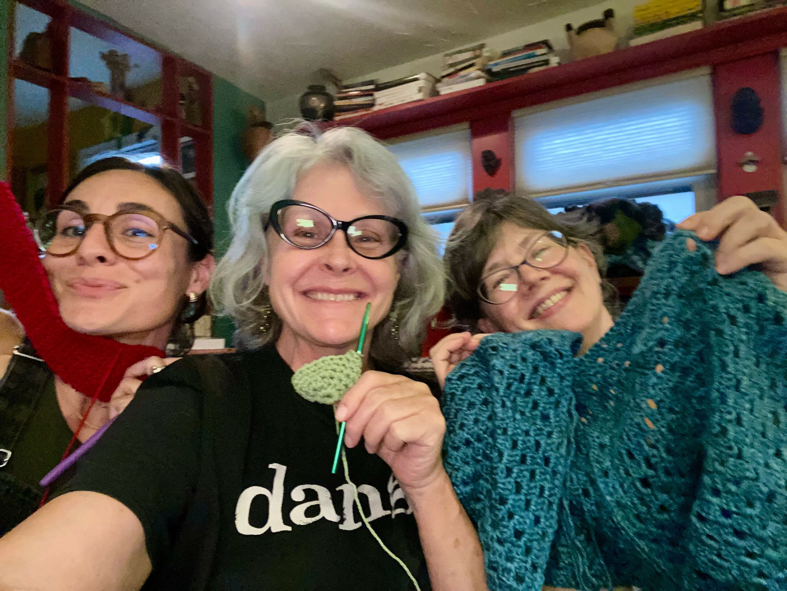 Three women smiling and posing indoors, one holding a green object, with colorful background and windows.