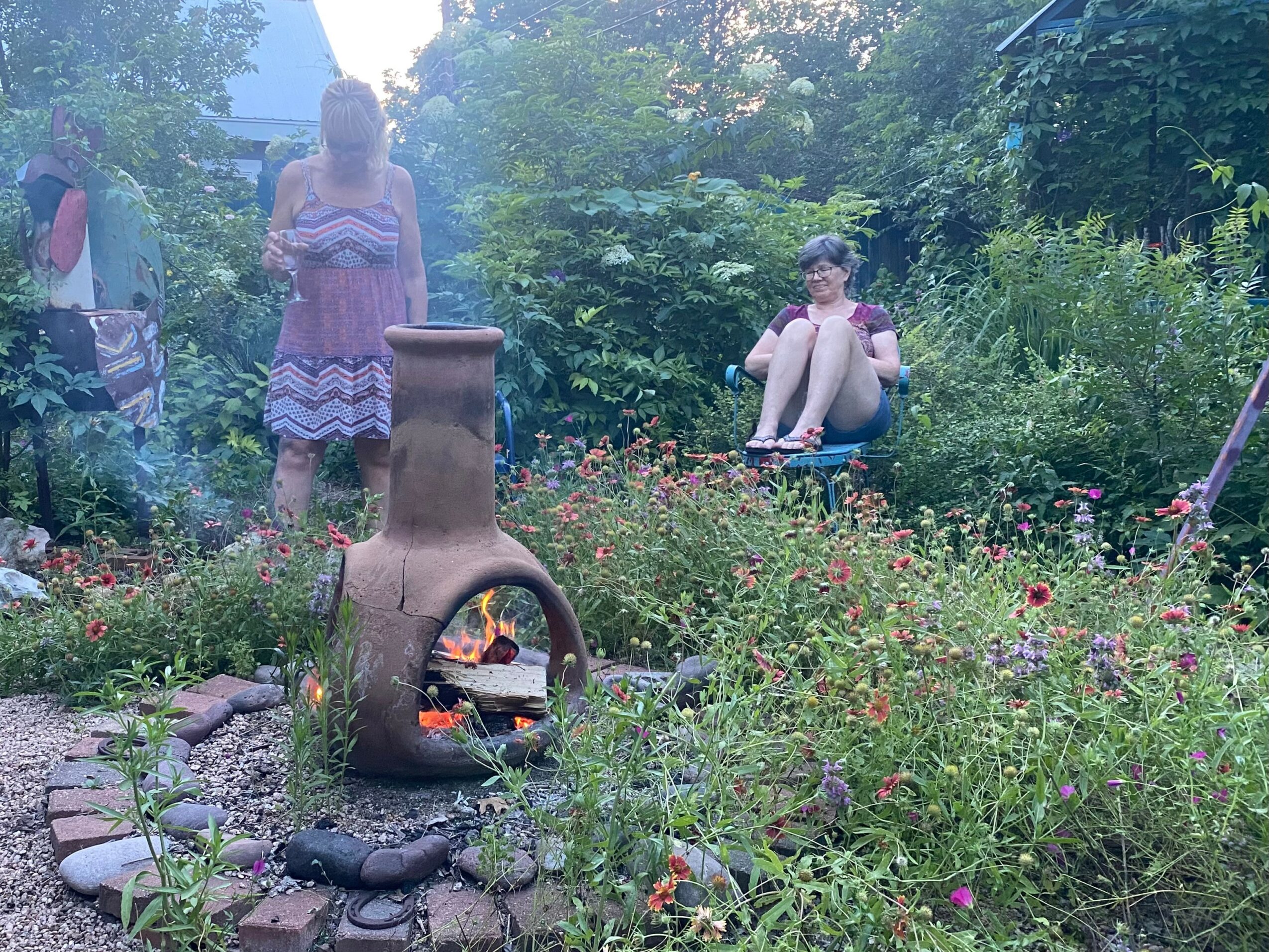 Two women in a garden with flowers and greenery, one standing and one sitting on a swing, near a chiminea.