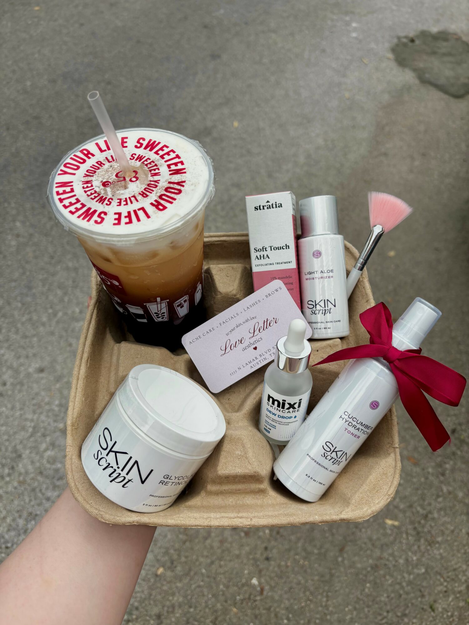 Hand holding a tray with skincare products, a drink, and a makeup brush on a concrete surface.