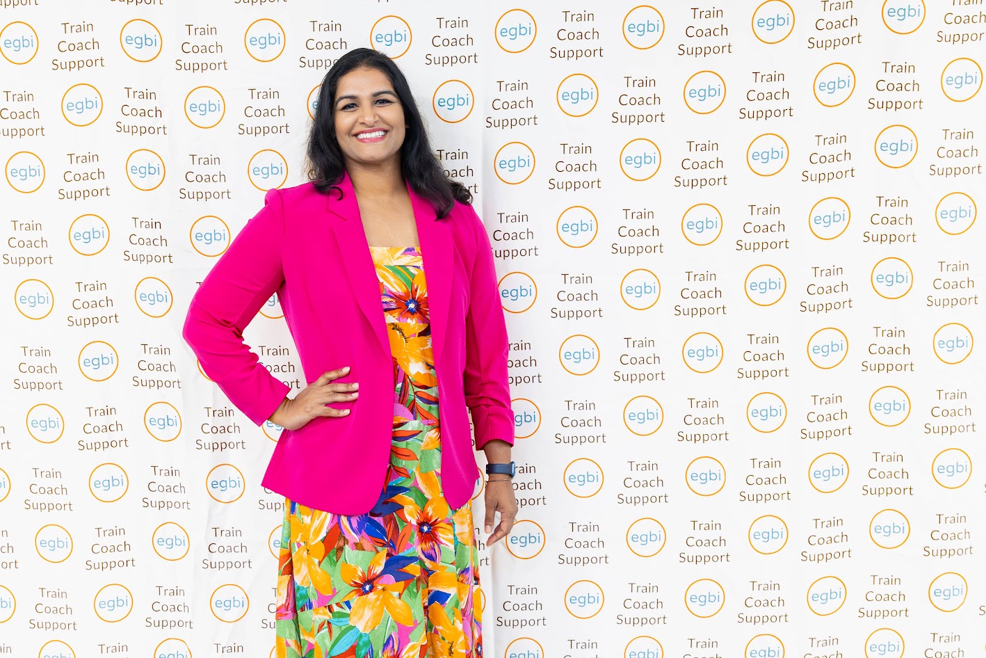 Woman in a bright pink blazer and colorful dress standing in front of a patterned backdrop with logos.