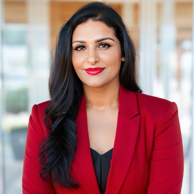 Woman with long black hair, red lipstick, wearing a red blazer and black top, smiling in a bright indoor setting.