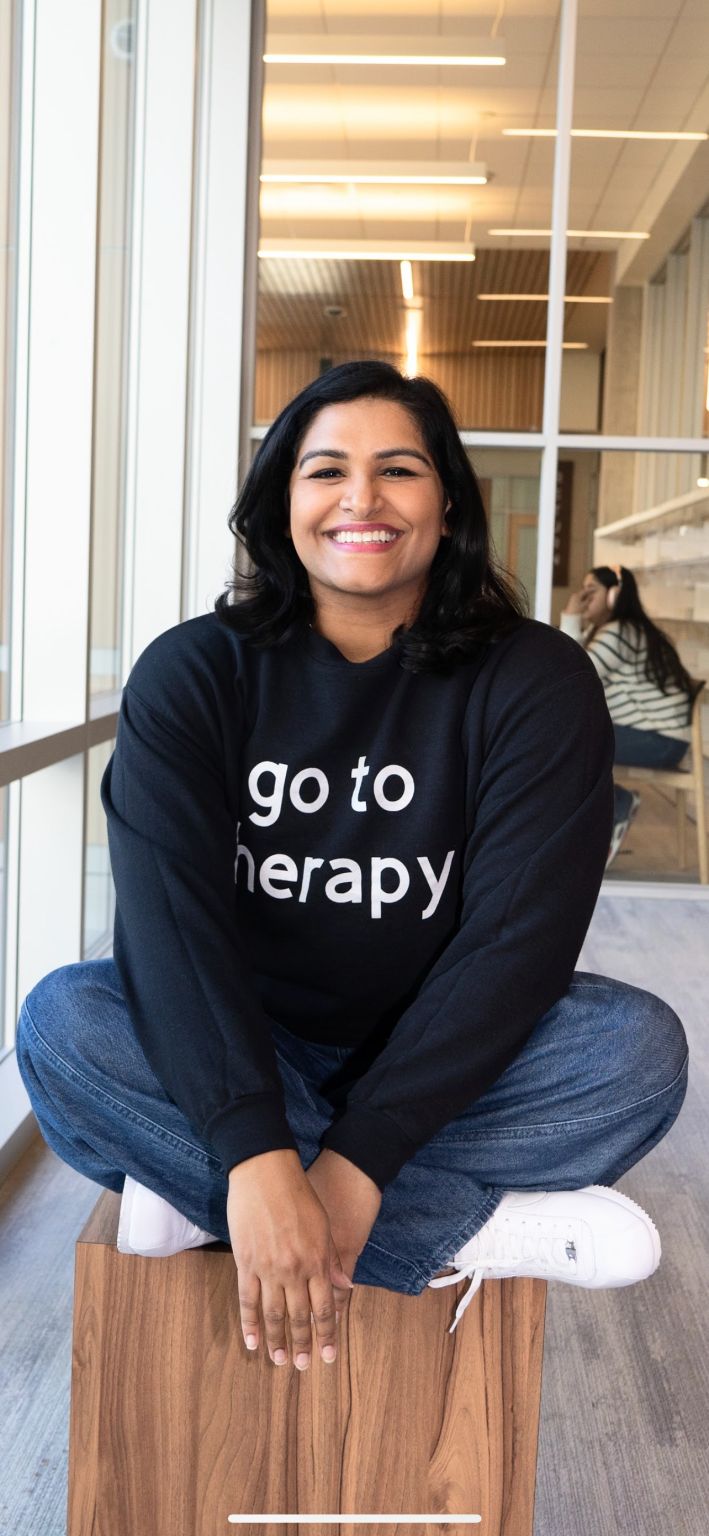 Smiling woman sitting cross-legged on a wooden block indoors, wearing a black sweatshirt with white text, in a modern space.