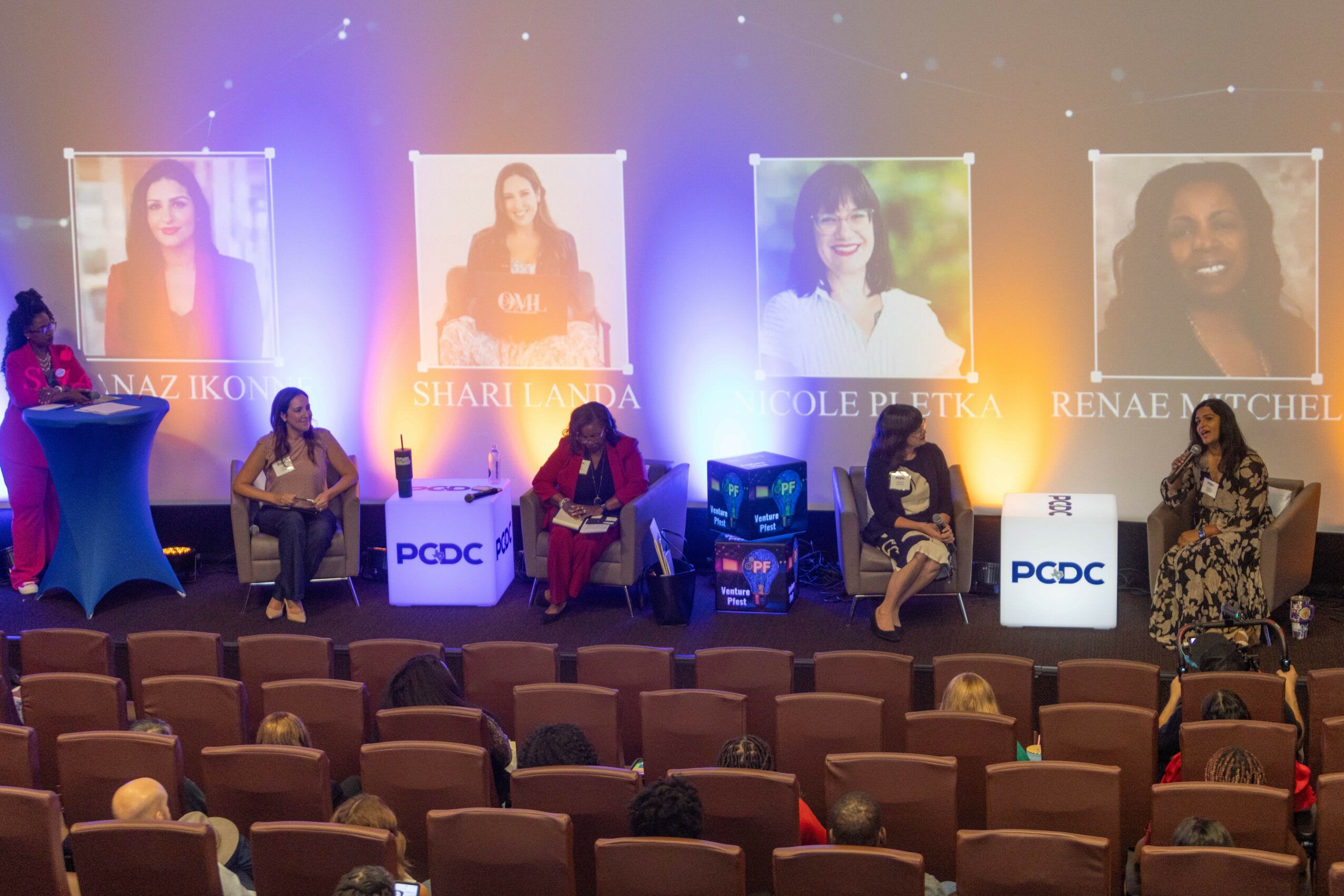 Six women seated on stage with large banners of their portraits behind them, audience seats in front, stage lit with purple and blue lights.