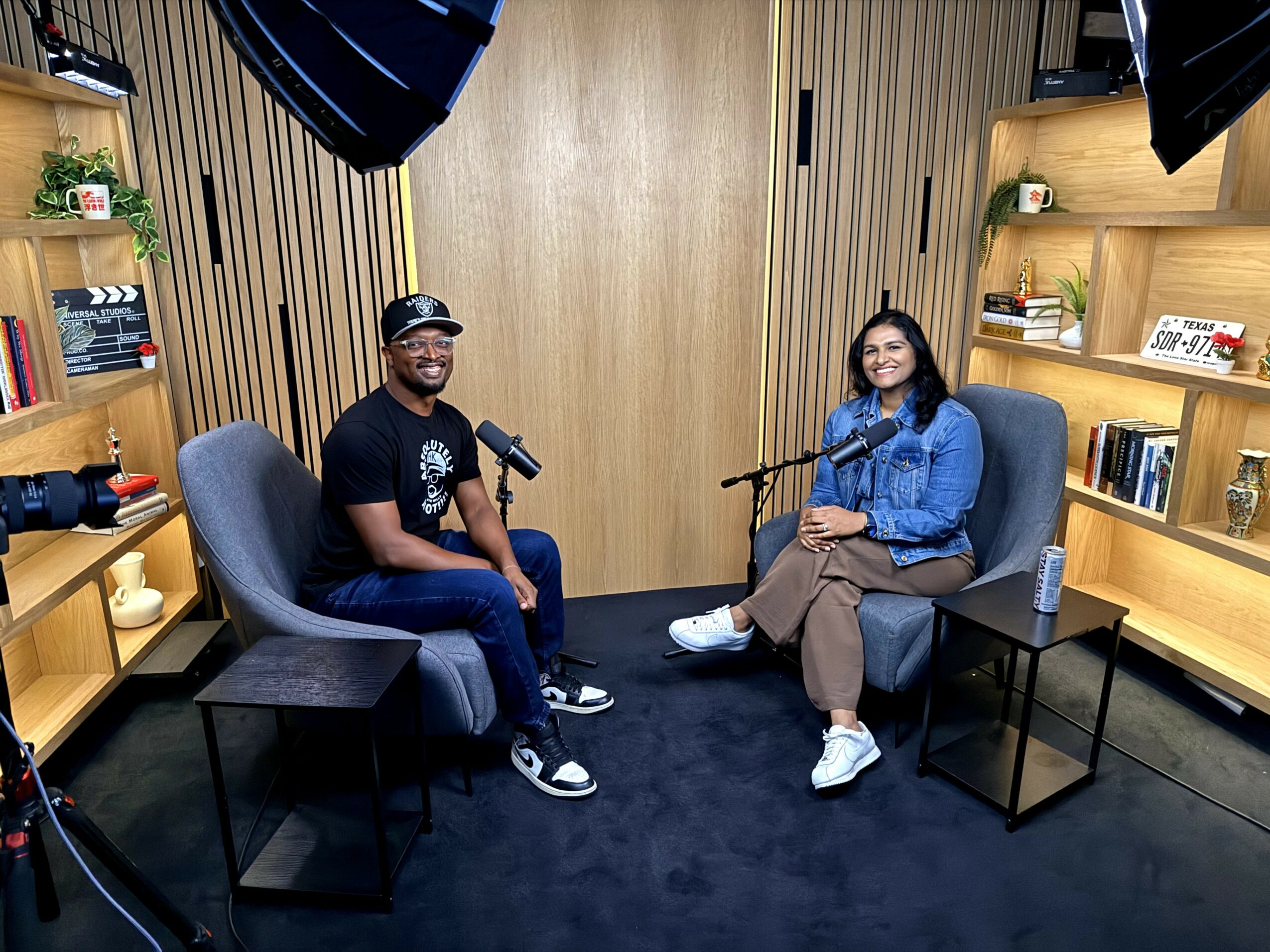 Two people sit in chairs with microphones in a room with wooden shelves and books, smiling at the camera.