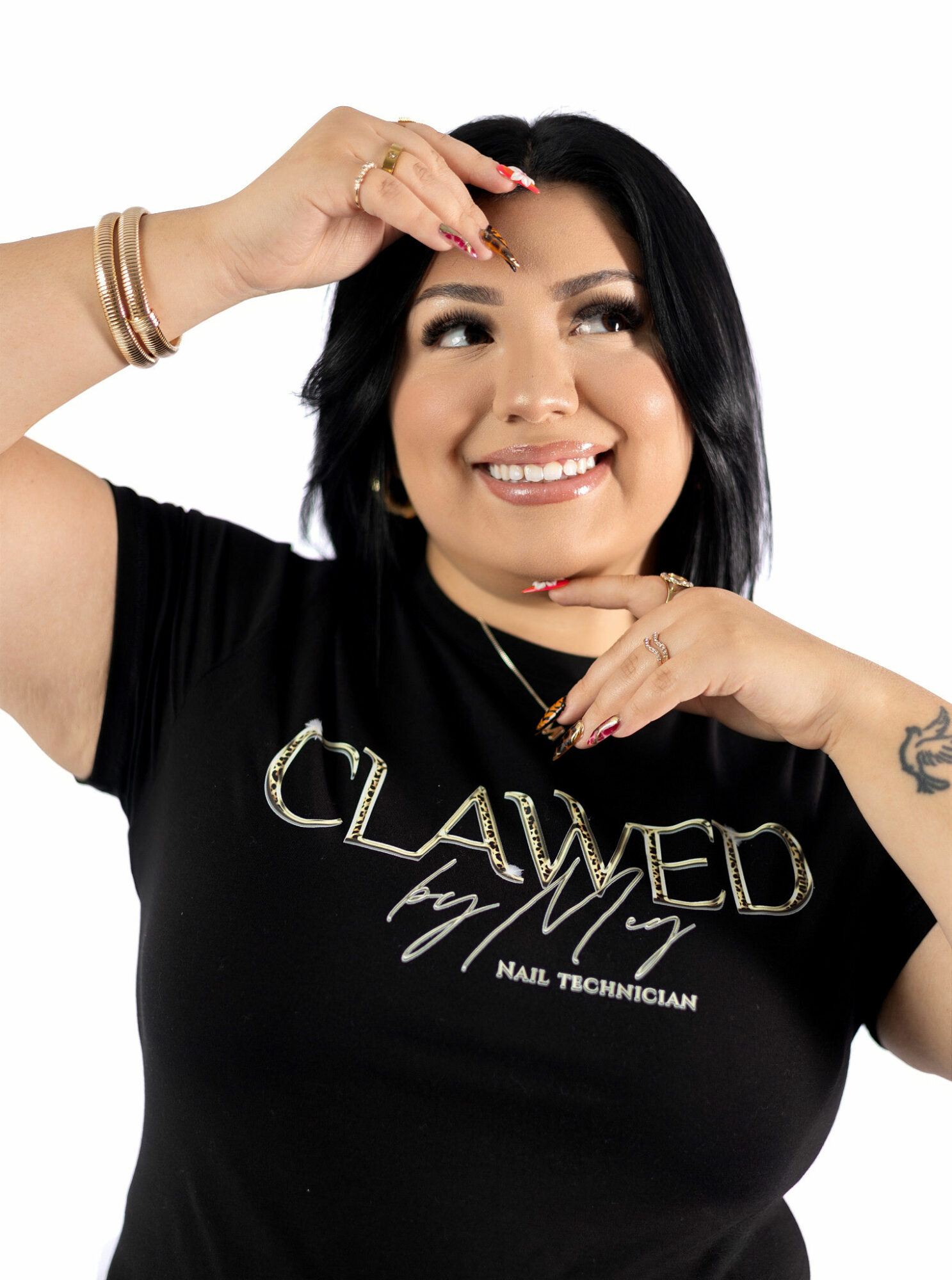 Smiling woman with black hair, wearing a black T-shirt with gold text, touching her forehead and chin, against a white background.