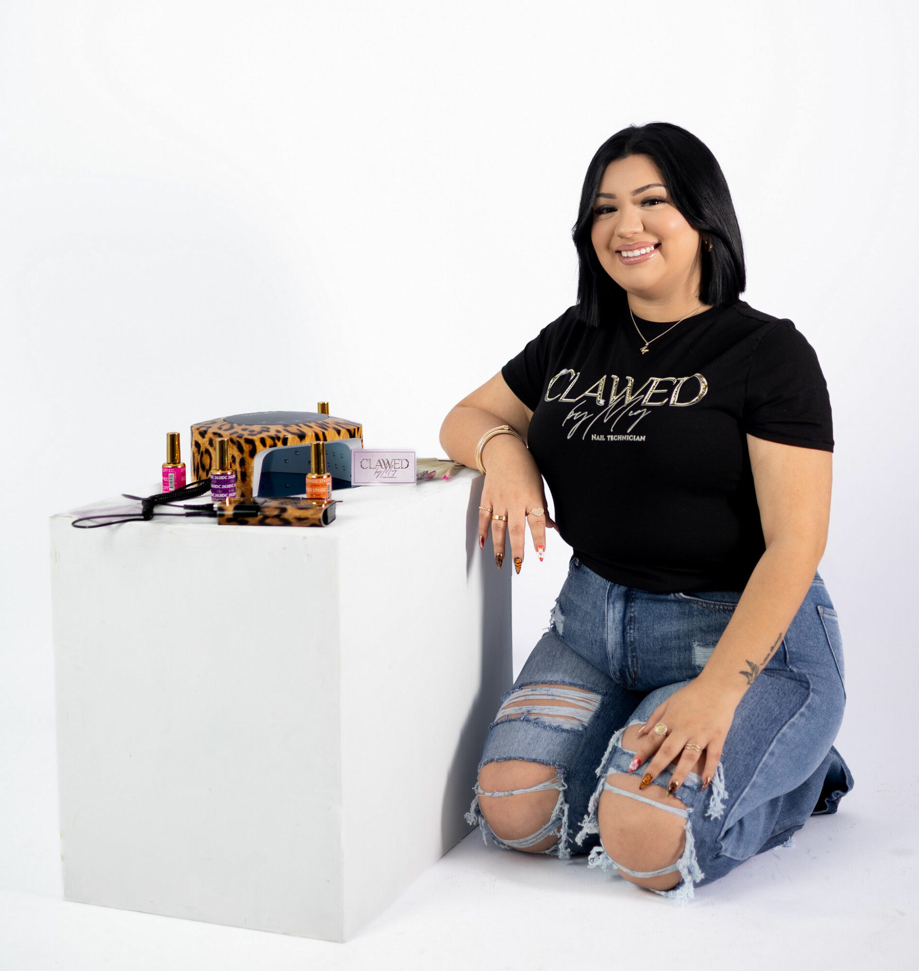 Woman kneeling and smiling next to a white display table with makeup items, wearing a black T-shirt and ripped jeans.