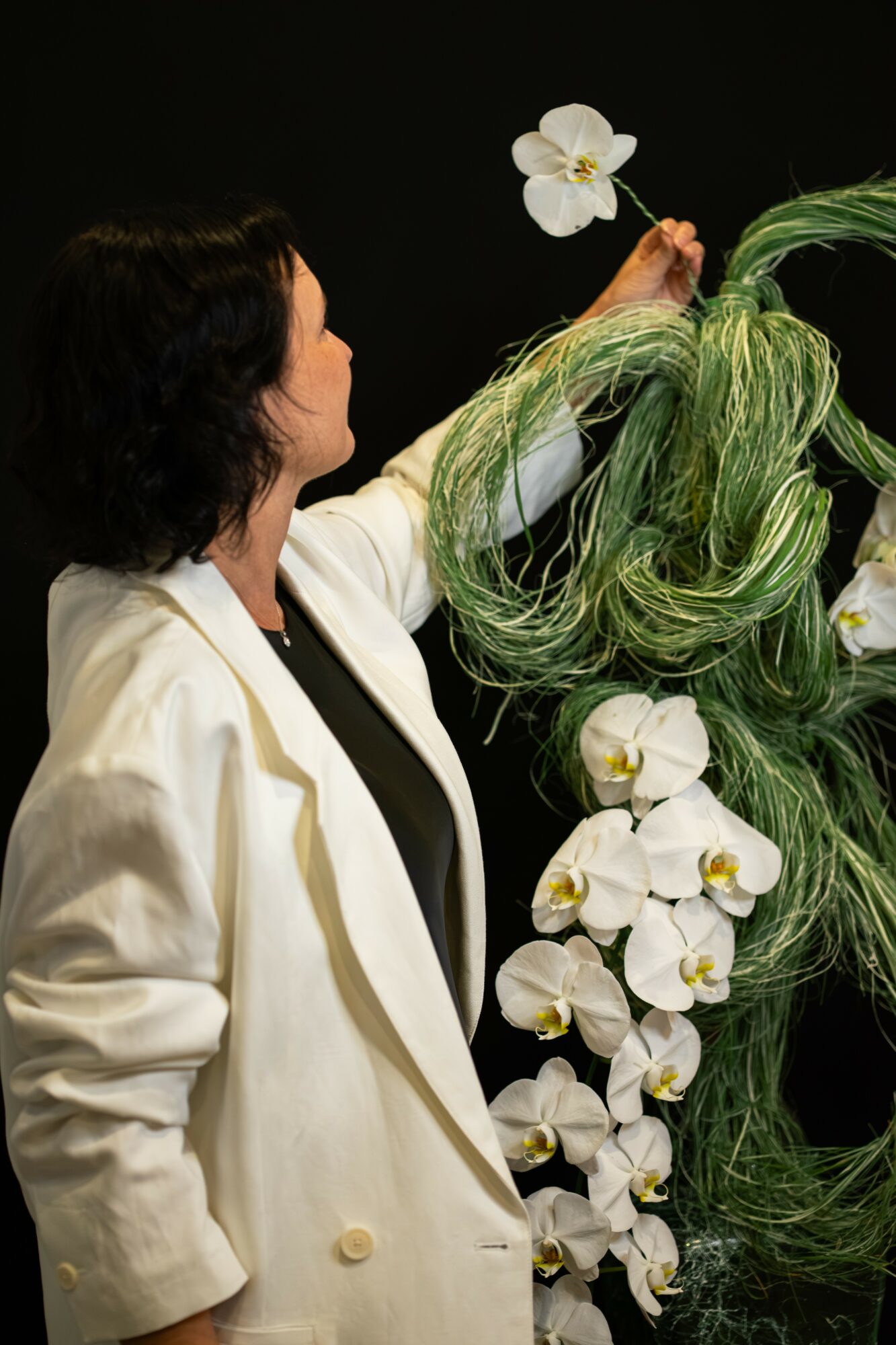 Woman with dark hair in a white blazer arranging white orchids and green foliage against a black background.