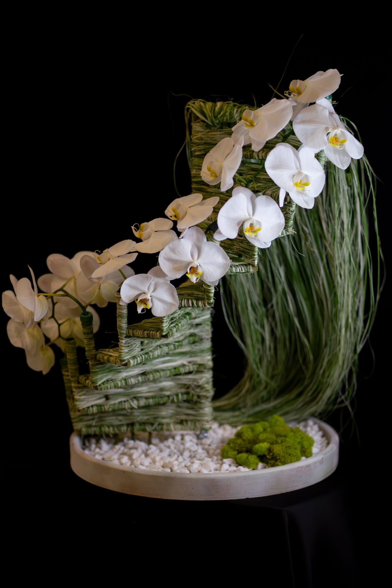 Arrangement of white orchids and greenery in a glass vase on a white base, with small white stones and green moss, against black background.