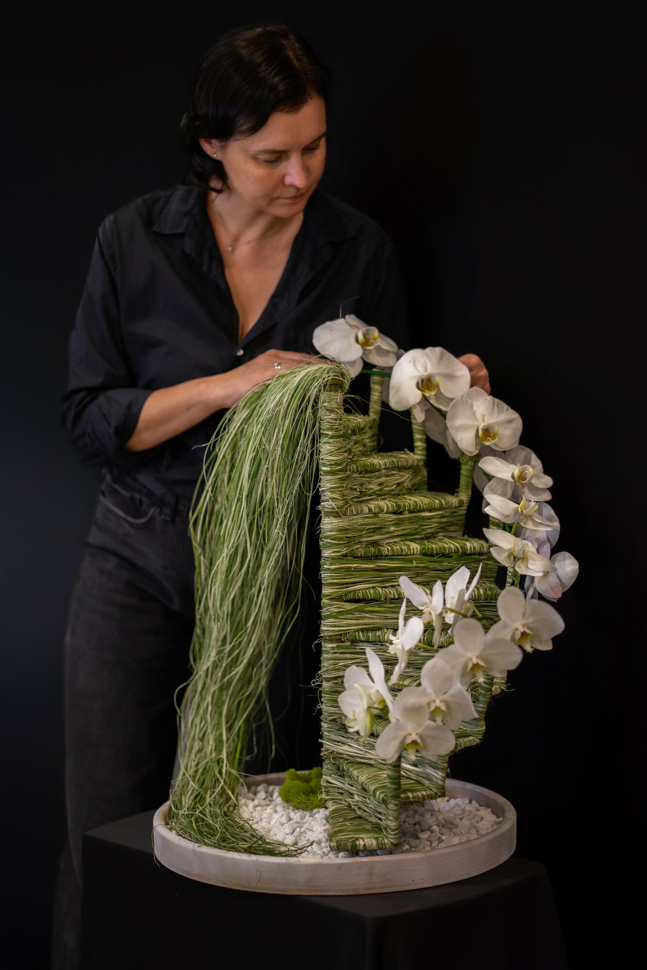 Woman arranging white orchids and green plants on a vertical display, black background, standing at a table.