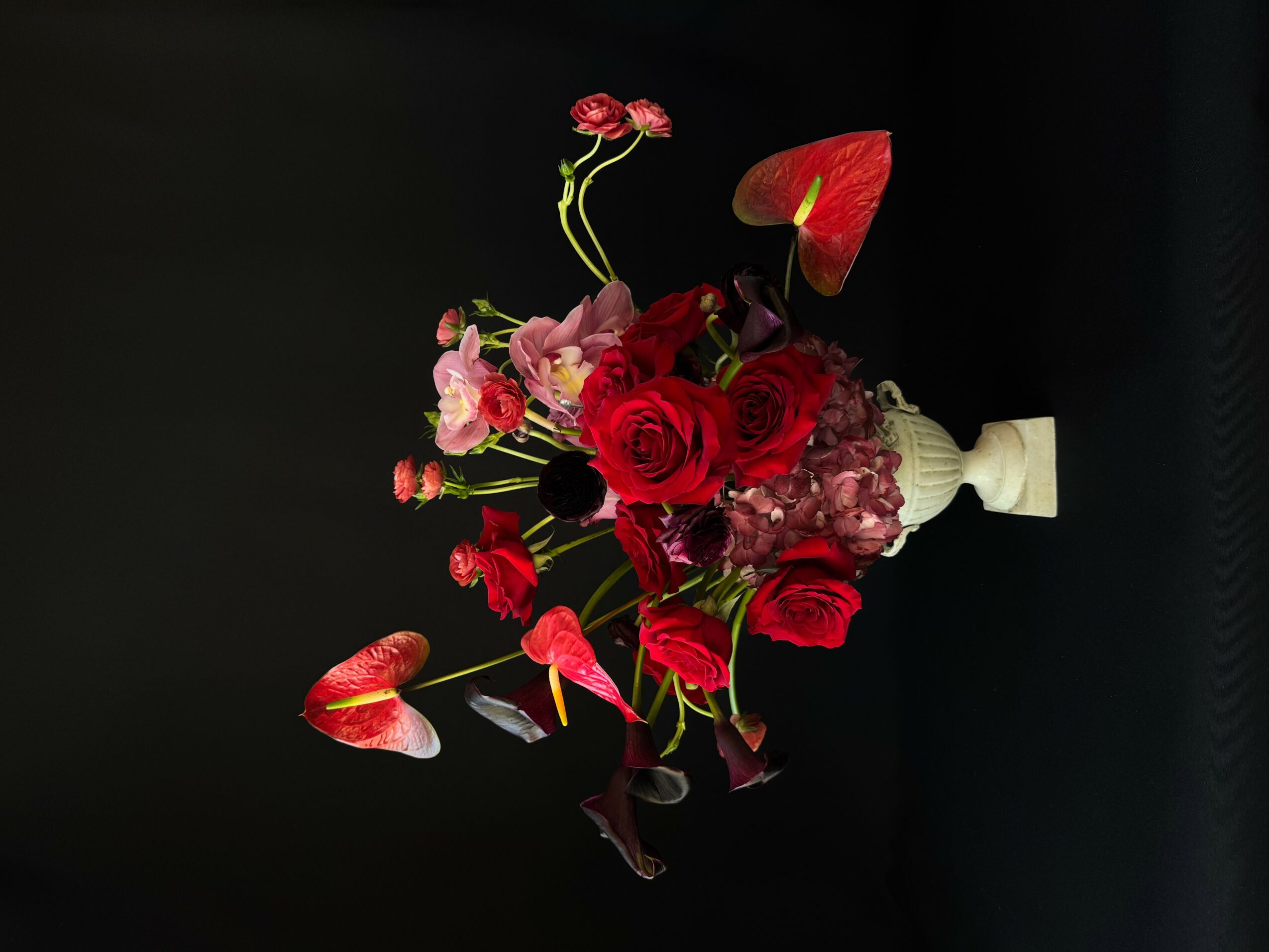 Flower bouquet with red roses, pink flowers, and red anthuriums in a white vase against a black background.