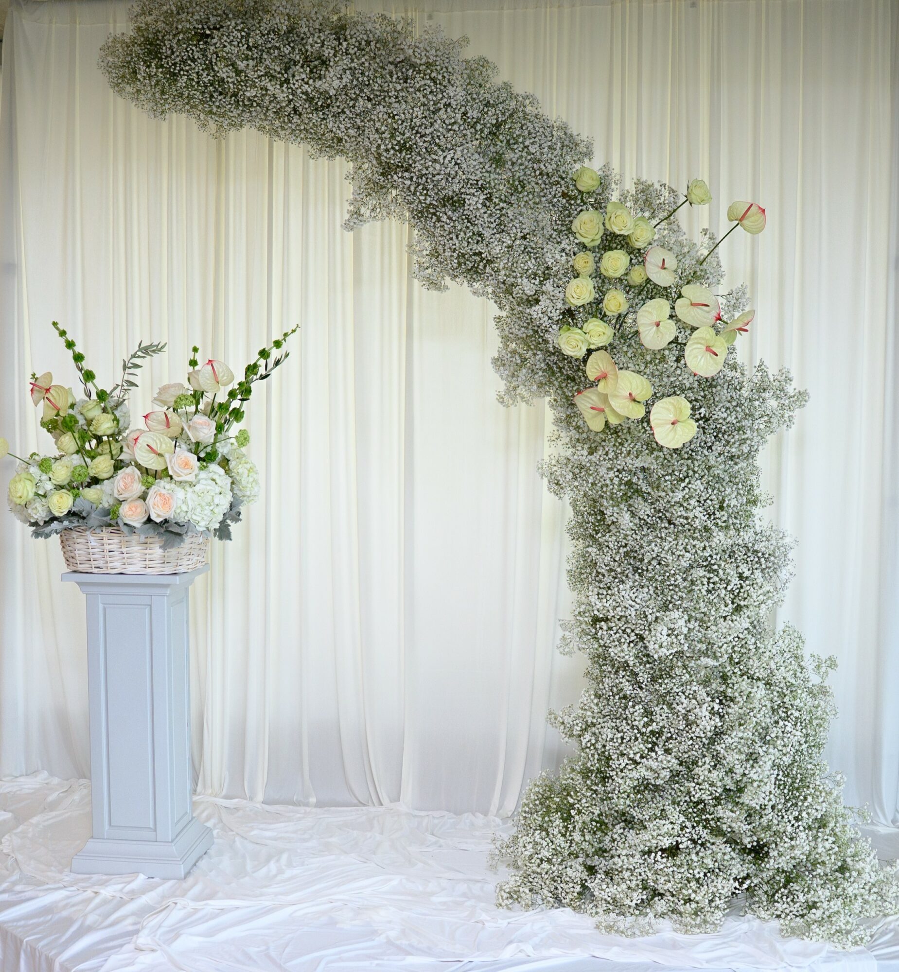 Floral arrangement with white and green flowers on a tall pedestal, with a curved floral arch against a white curtain backdrop.