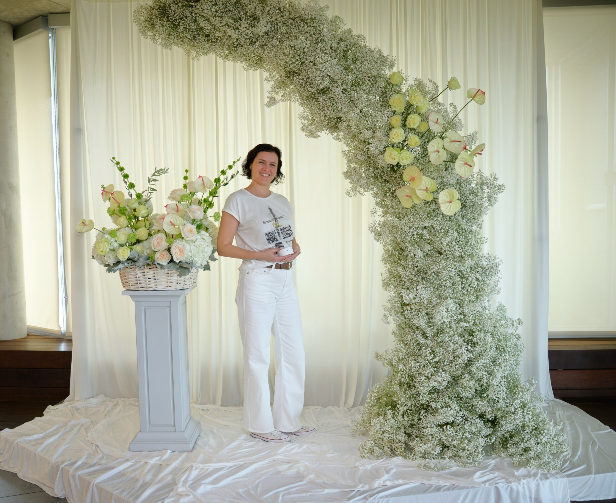 Woman standing next to a floral arrangement on a pedestal, with a large floral arch behind her, in a decorated indoor setting.