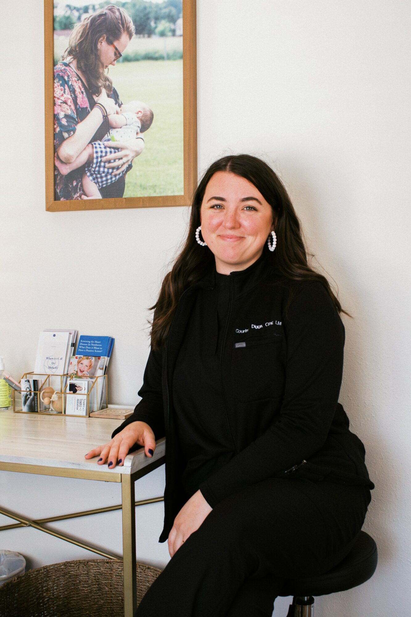 Woman with long dark hair sitting on a stool, smiling, in a room with a framed photo on the wall behind her.