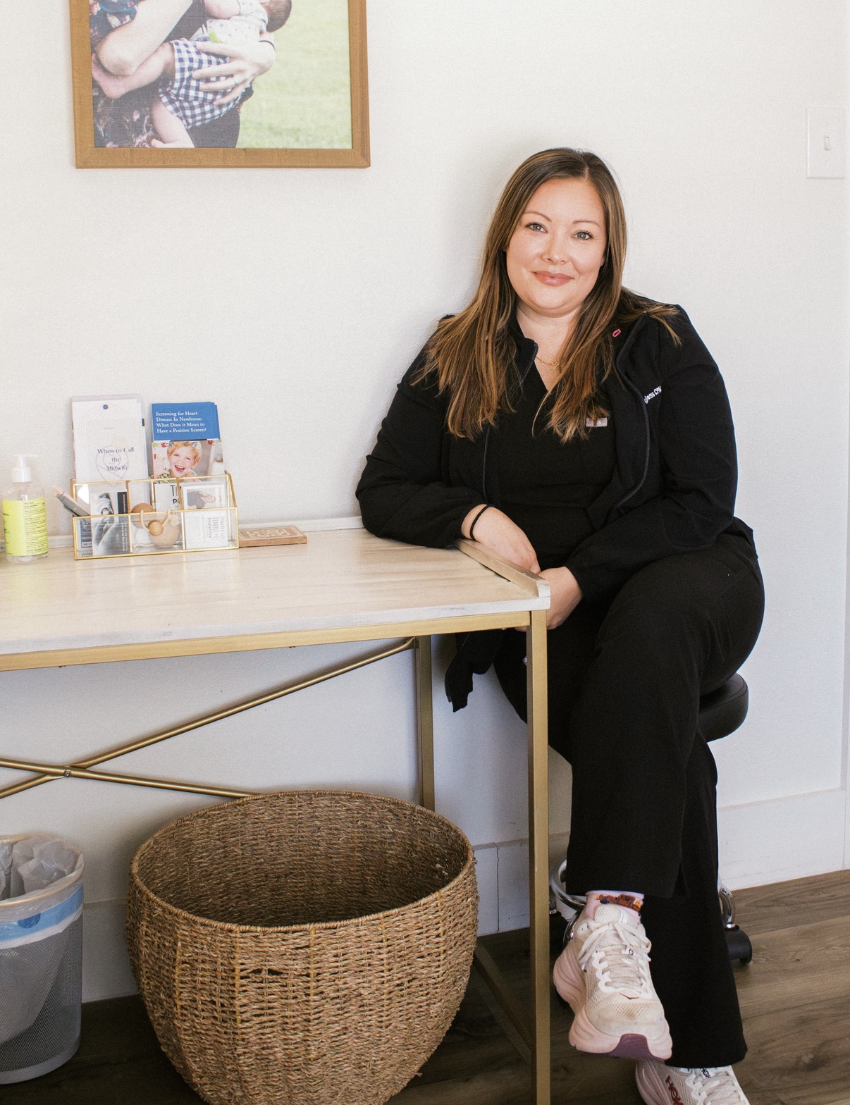 Woman sitting at a desk with a smile, in a room with a framed photo on the wall, a basket below, and a white wall.