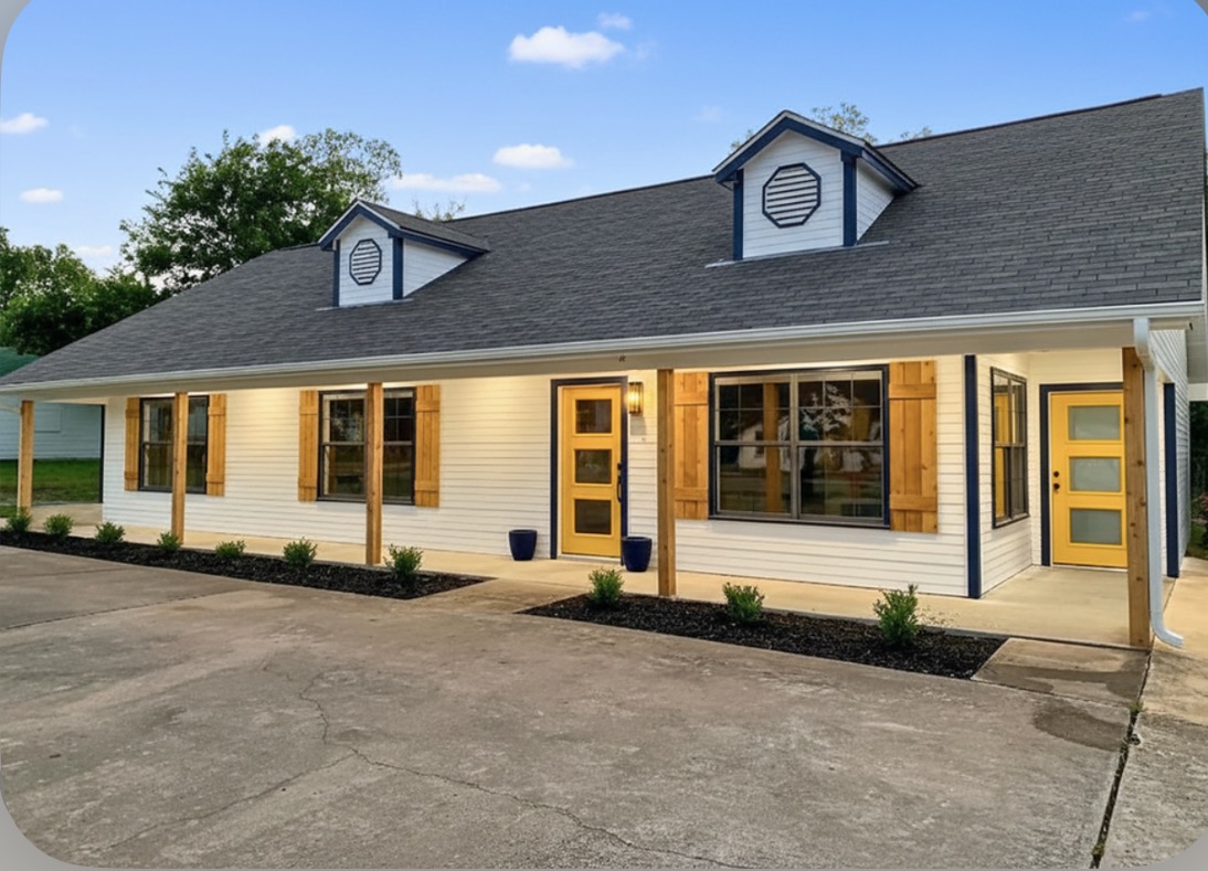 Single-story building with yellow doors, large windows, and a porch with wooden posts, under a gray roof with dormer windows.