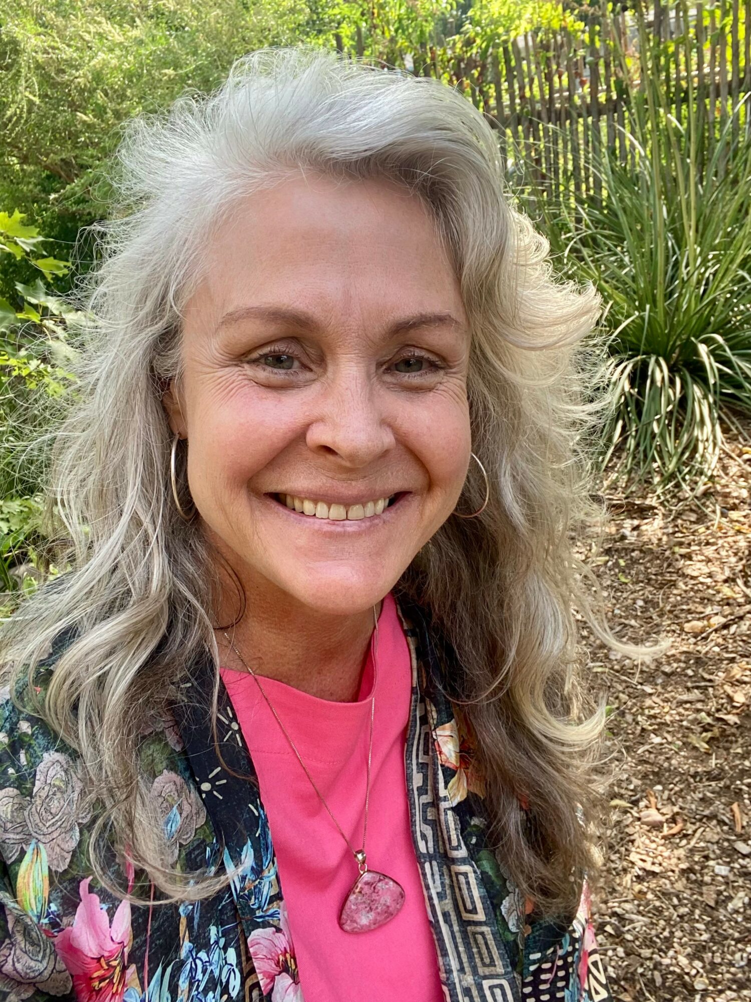 Smiling woman with long, curly gray hair outdoors, wearing a pink shirt, patterned jacket, and a heart-shaped pendant.